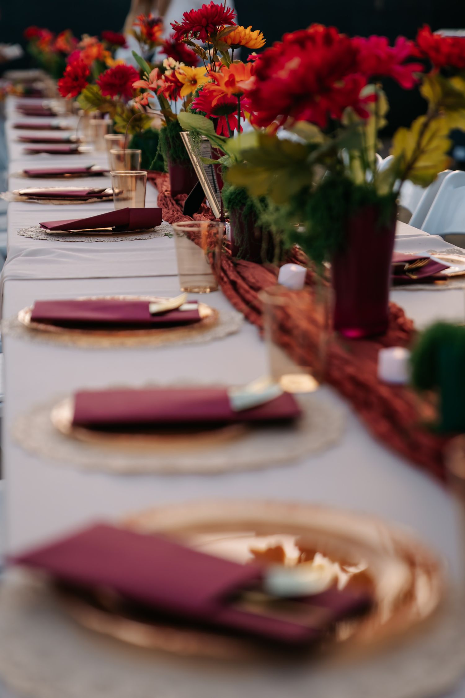 Elegant fall wedding table setting with burgundy napkins, gold charger plates, and red floral centerpieces on a white tablecloth.