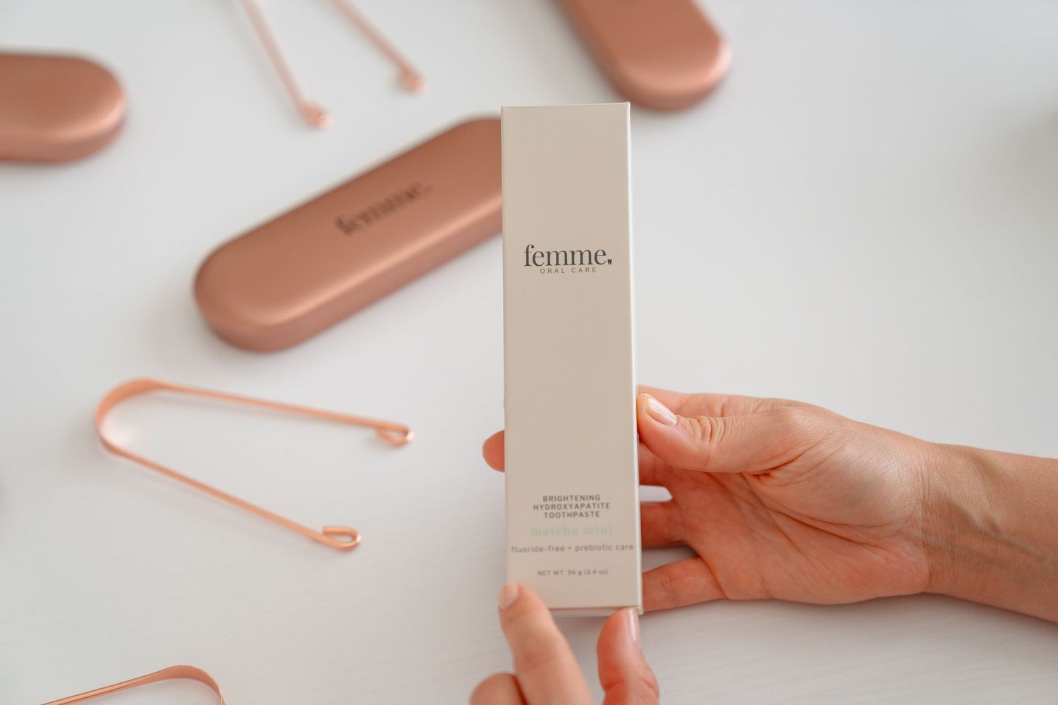 Hands holding a Femme branded skincare product box with rose gold accessories on a white background.
