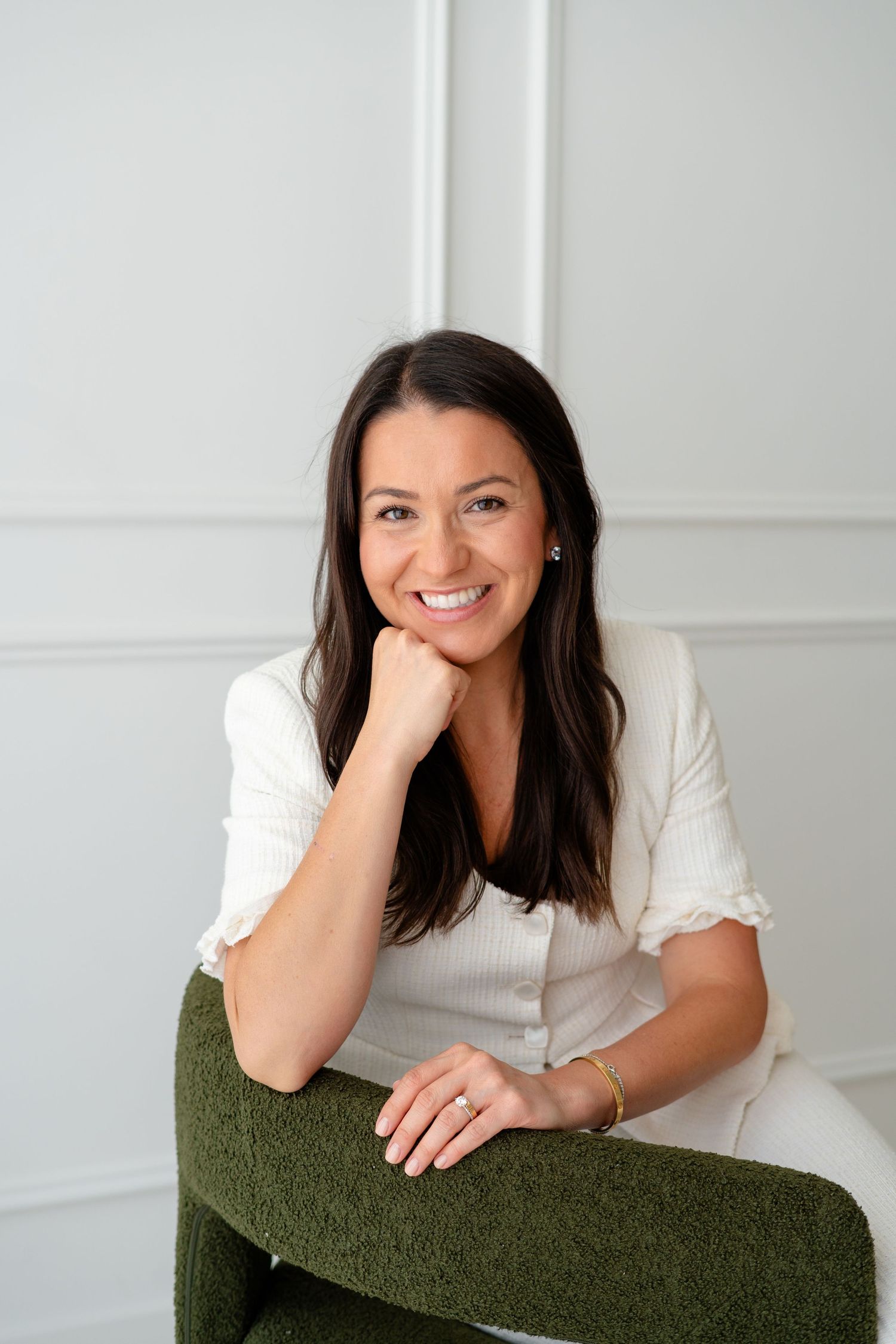 Smiling brunette woman in white top and green pants sitting relaxed with hand on chin against white wall.
