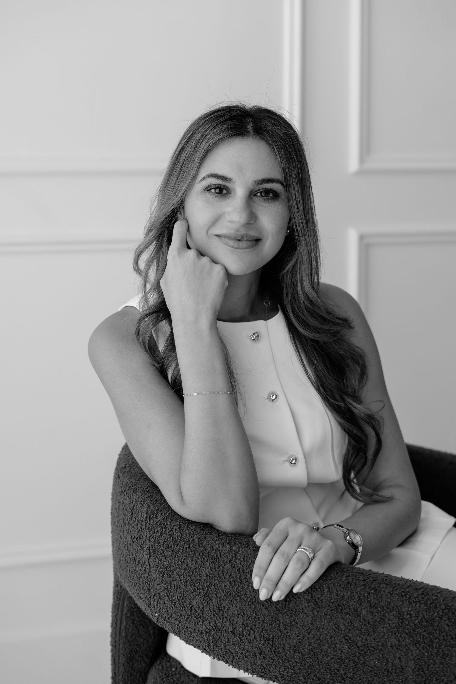 Black and white portrait of a smiling woman with long wavy hair, seated in a chair, wearing a sleeveless top.