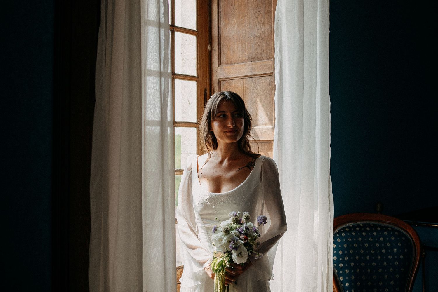 Bride in white gown holding wildflower bouquet stands by sheer curtained window in elegant dark-walled room.