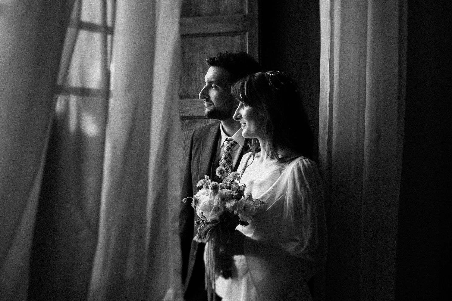 Black and white wedding photo of bride holding bouquet and groom gazing out window with sheer curtains.