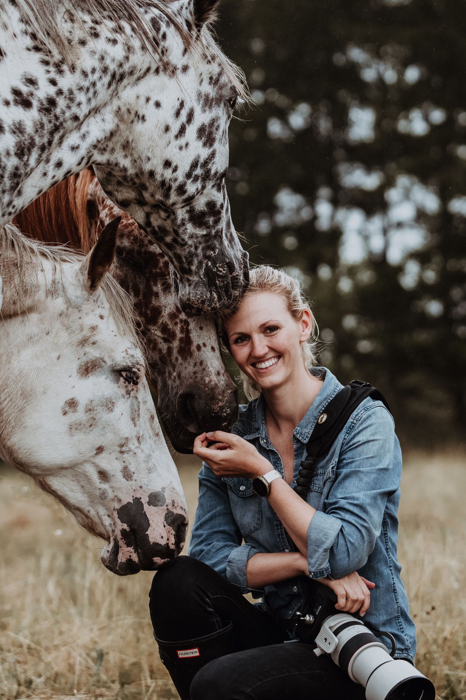 Smiling woman photographer crouching with camera beside two spotted Appaloosa horses in a field.