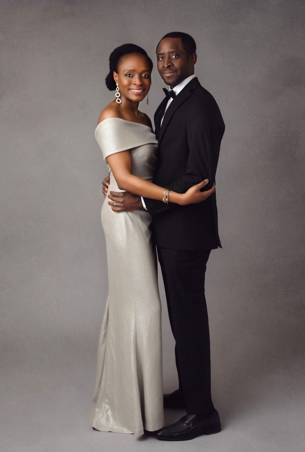 Elegant couple posing in formal attire, woman in silver gown and man in black tuxedo, studio portrait.