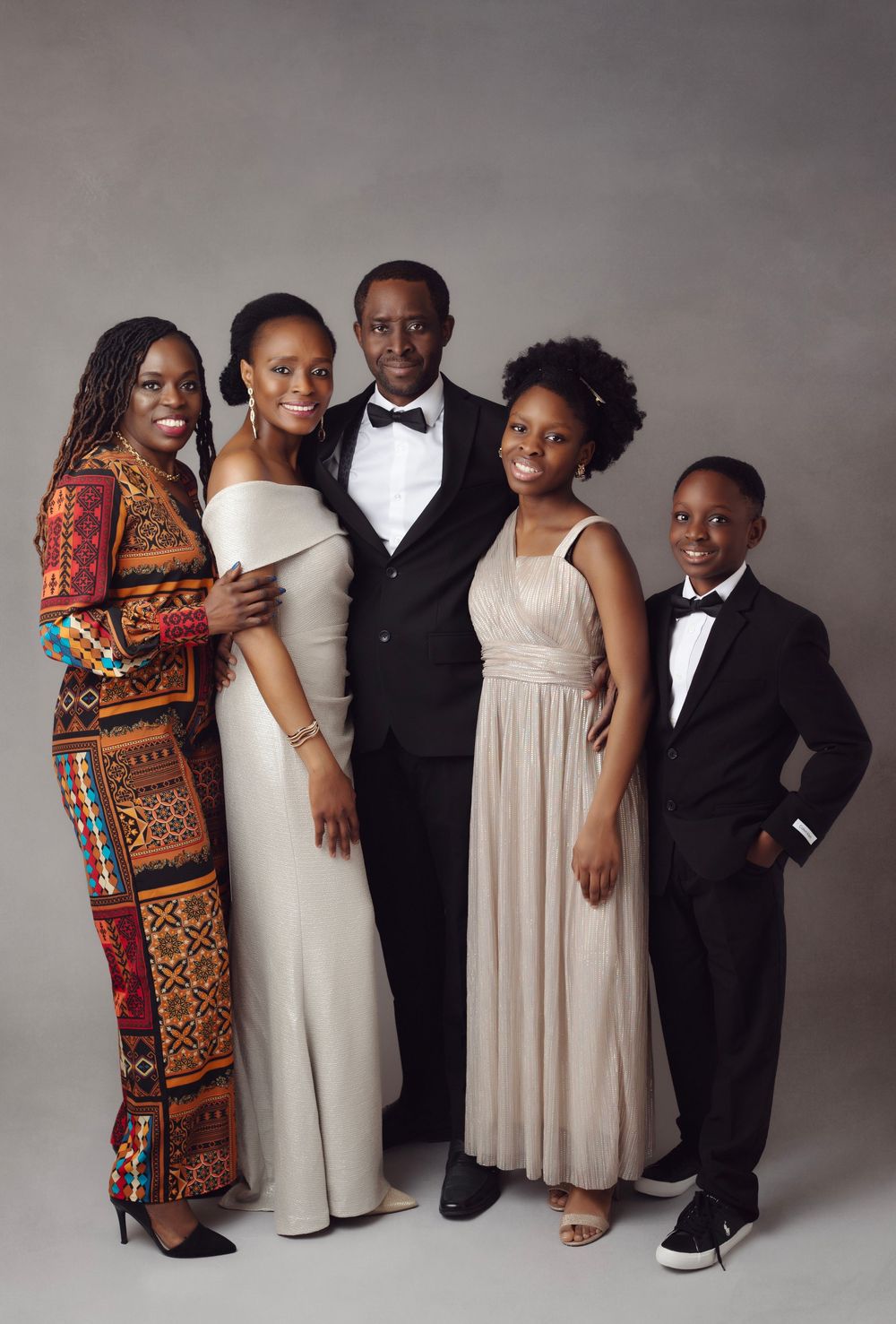 A joyful family of five poses in formal attire, including elegant gowns and tuxedos, against a gray studio backdrop.