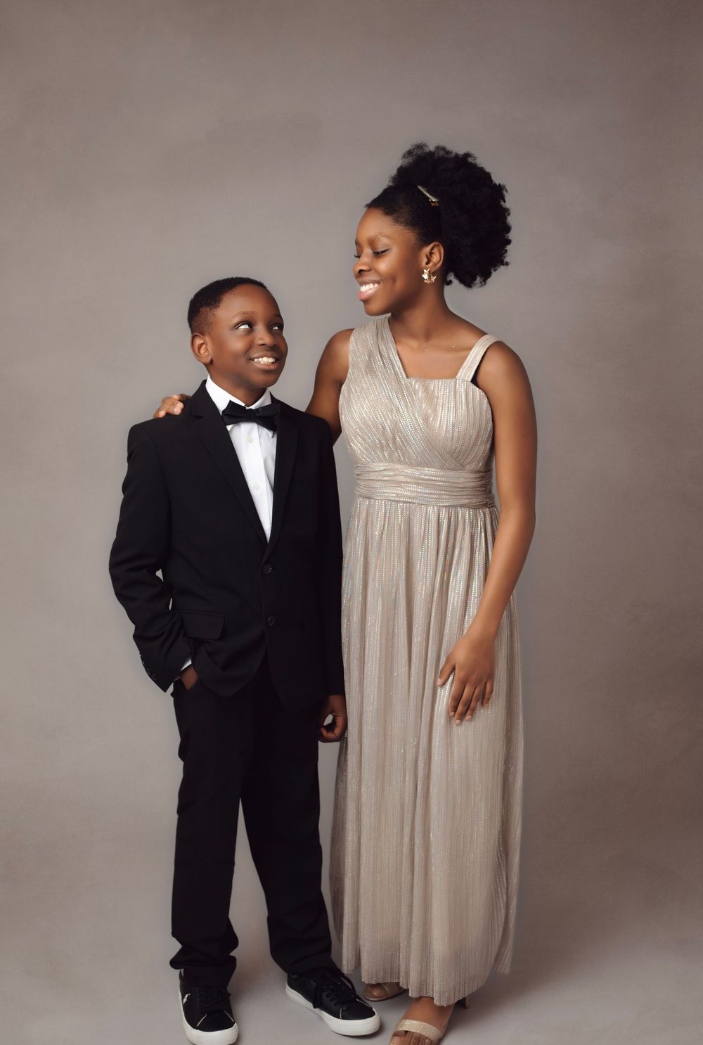 A young boy in a tuxedo and a teenage girl in a silver gown smile together in a professional studio portrait.
