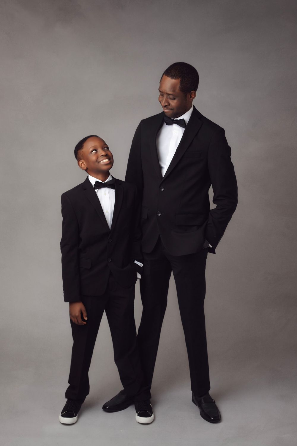Father and son duo in matching black tuxedos with bow ties posing together in studio.