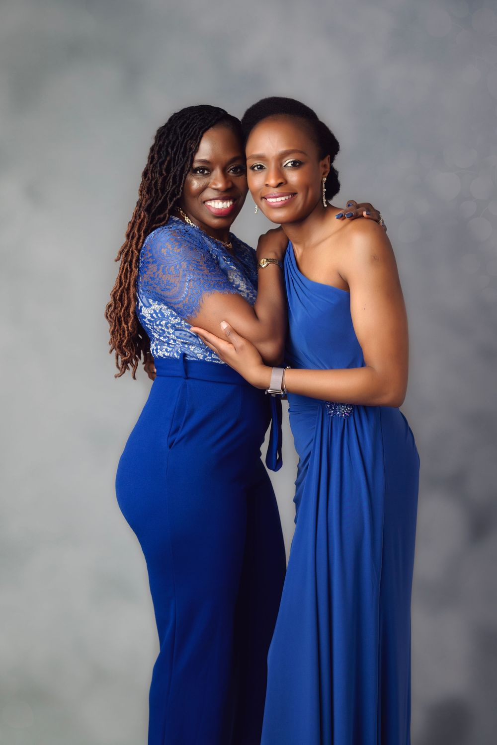 Two women in matching royal blue formal outfits smile and pose together against a gray studio background.
