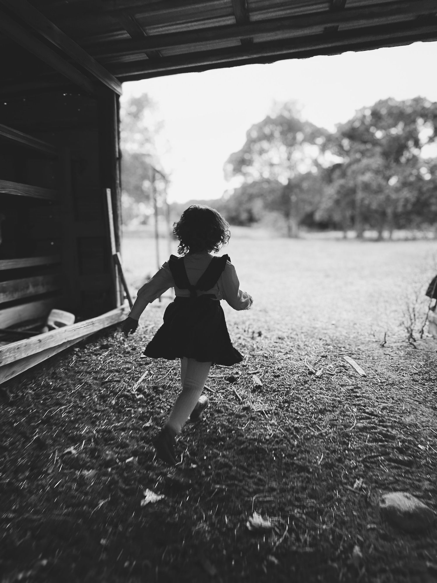 Black and white photo of a young curly-haired girl in a dress running toward an open field.