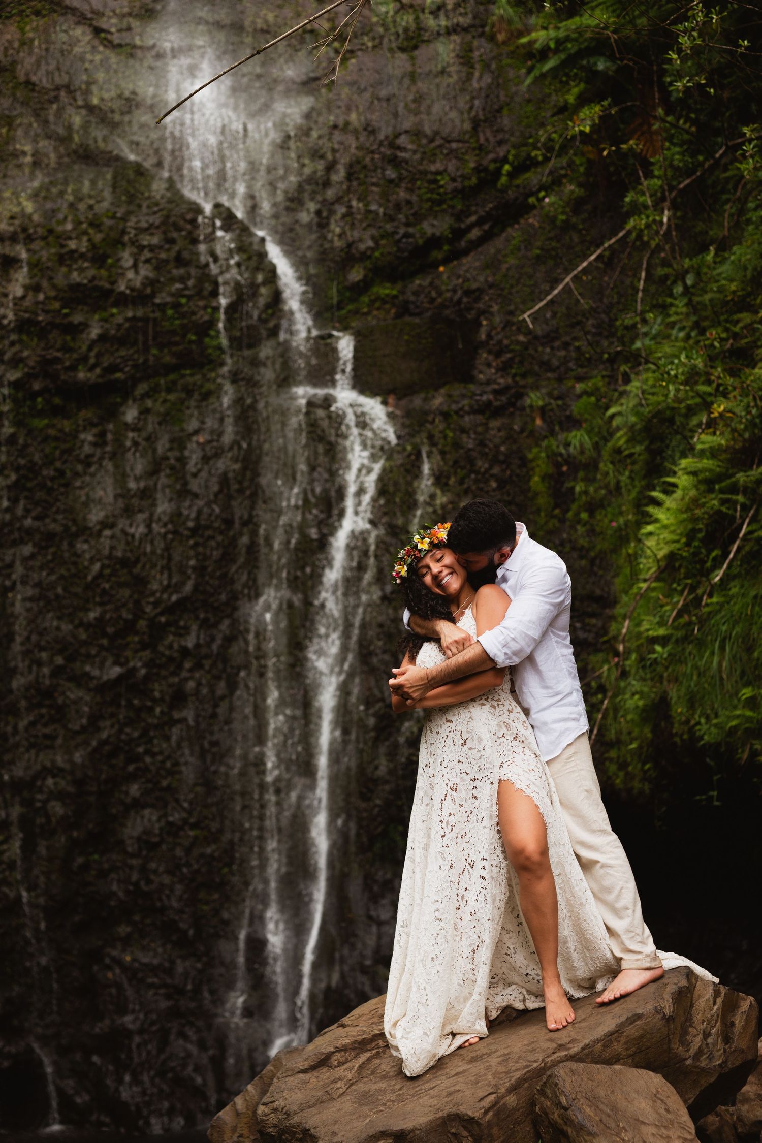 Couple embracing romantically in front of a lush waterfall, bride in white lace gown with floral crown. Maui Hawaii Elopement Photography.