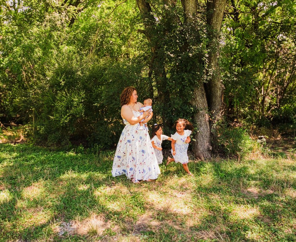A mother in a floral dress holds a baby while two young children play outdoors in a sunlit green forest setting.