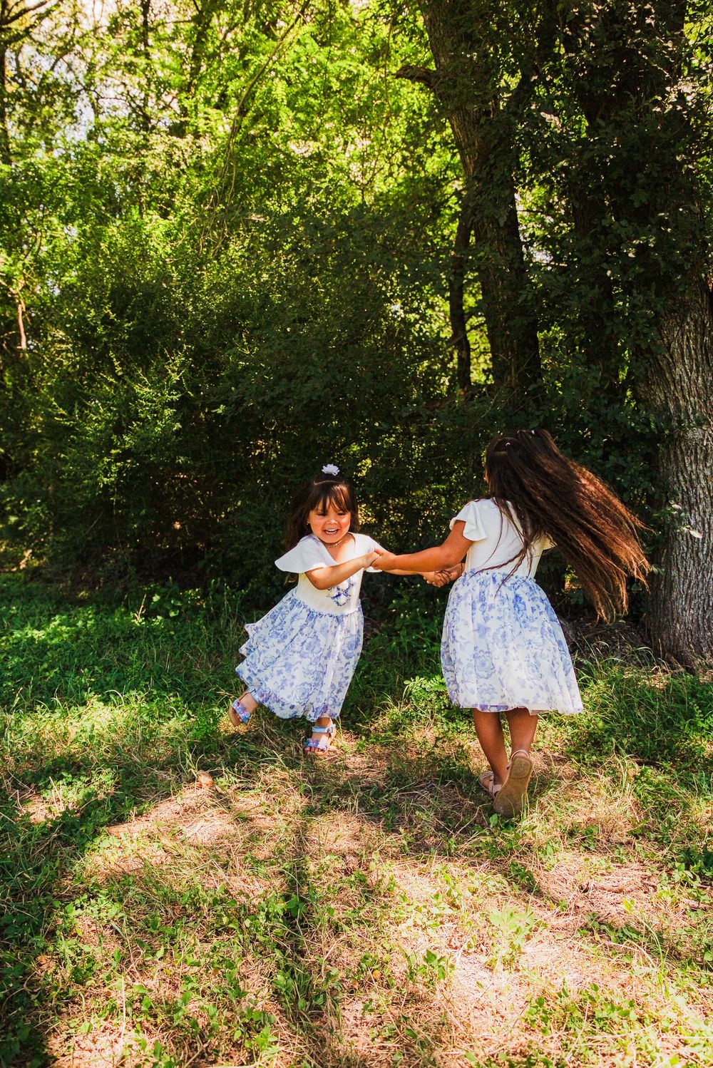 Two young girls in matching blue and white dresses hold hands and spin together in a sunny forest clearing.