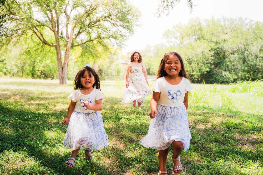 Three young girls in white dresses running and laughing through a sunny green park with trees in the background.