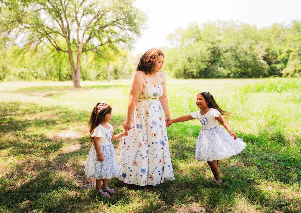 A mother in a floral dress holds hands with her two young daughters in matching white dresses in a sunny park.