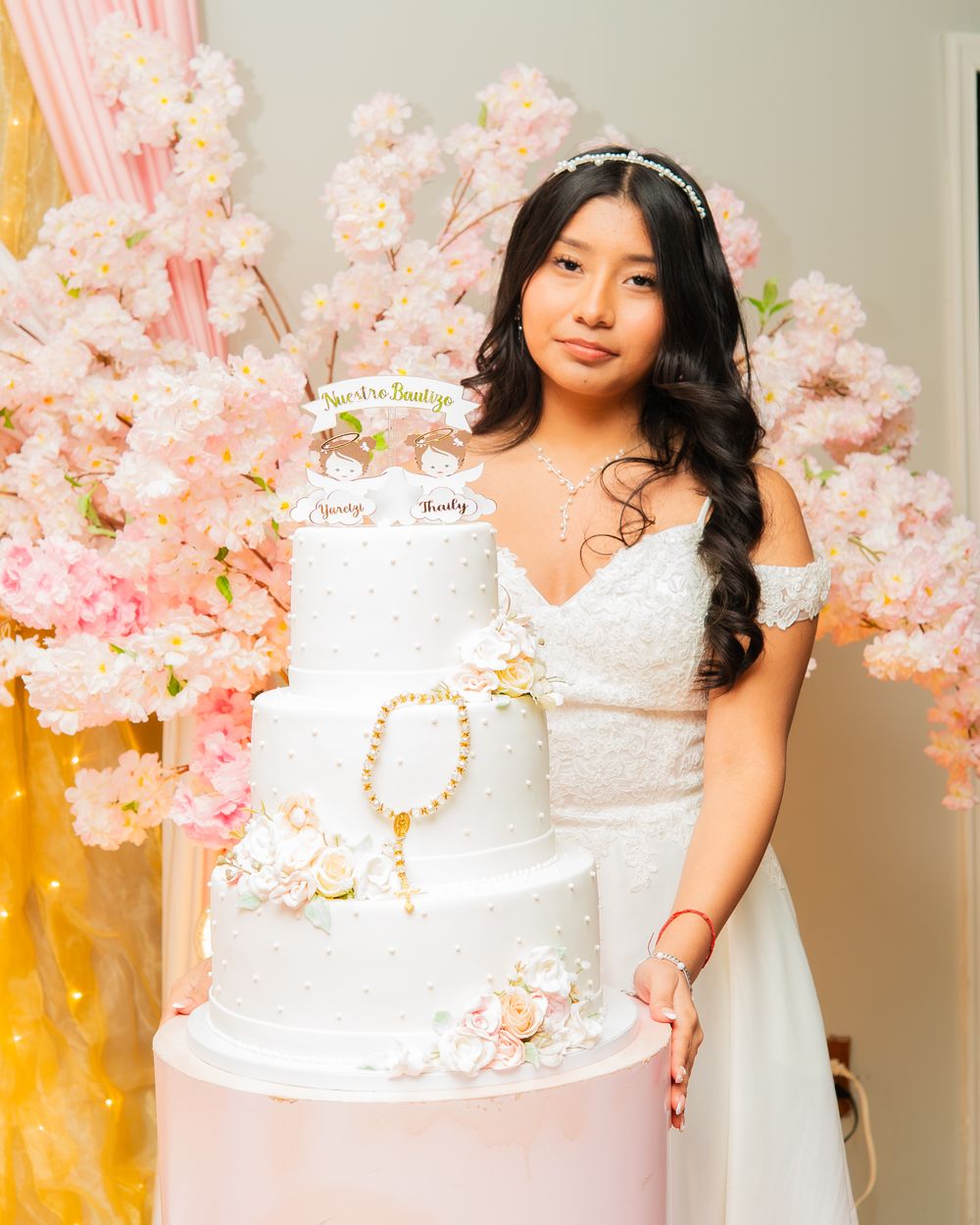 Young woman in white off-shoulder dress posing with elegant 4-tier white wedding cake adorned with pink flowers.