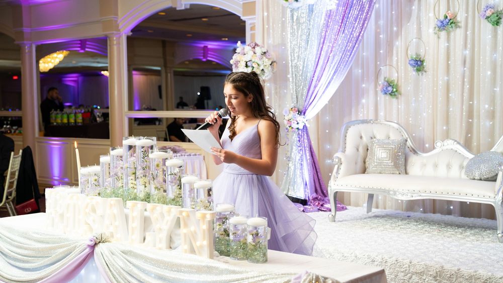 Young woman in lavender gown speaking at a quinceañera celebration with elegant white and purple decor.
