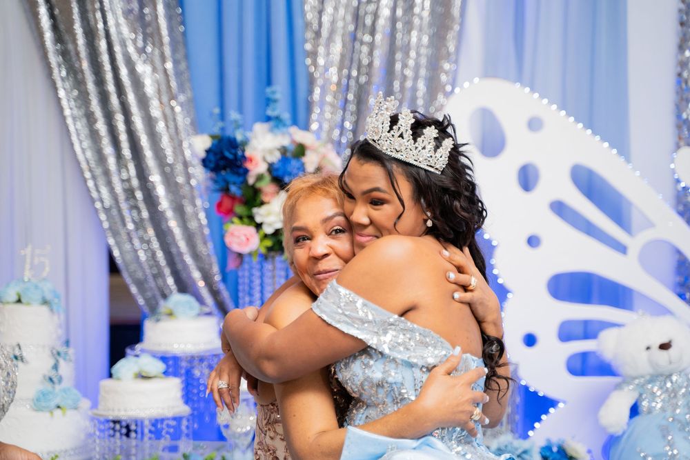 Two women embrace warmly at a quinceañera celebration, one wearing a tiara and blue gown near a decorated cake.