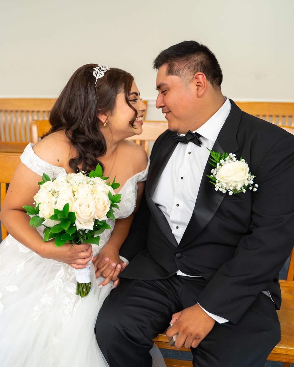 Bride and groom share a romantic moment on a church pew, bride holding white roses bouquet, groom in black tuxedo.
