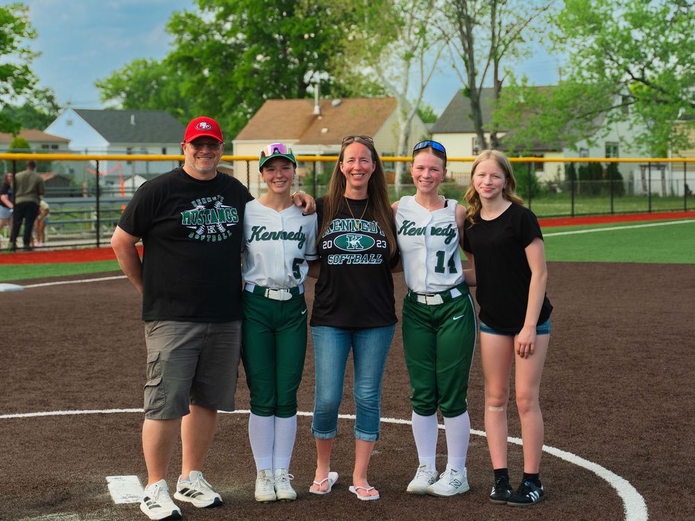 Group of five people posing on a softball field, with two players in green Kennedy uniforms during a sunny game day.