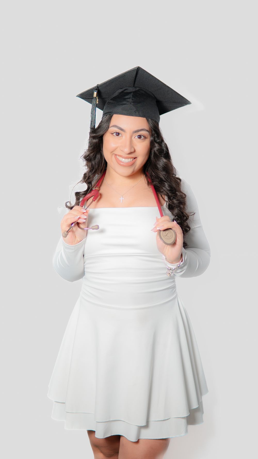 Smiling young woman in white dress and graduation cap holding a stethoscope against a light gray background.