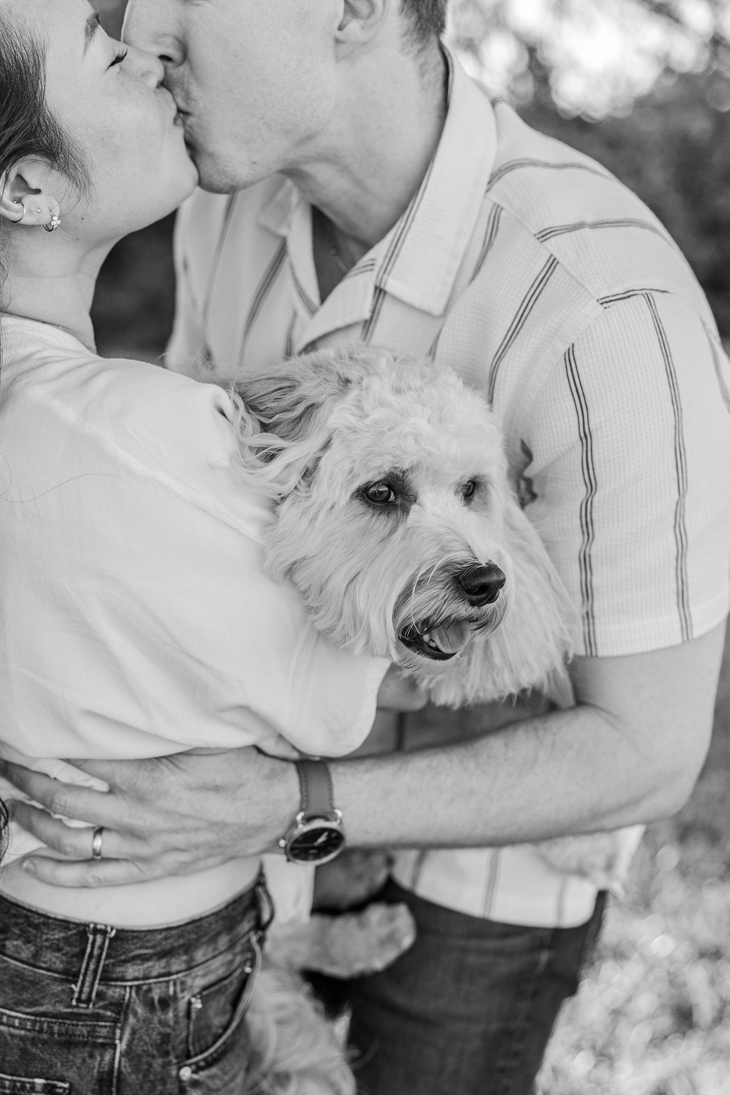 Black and white photo of a couple kissing while holding a fluffy white dog outdoors.