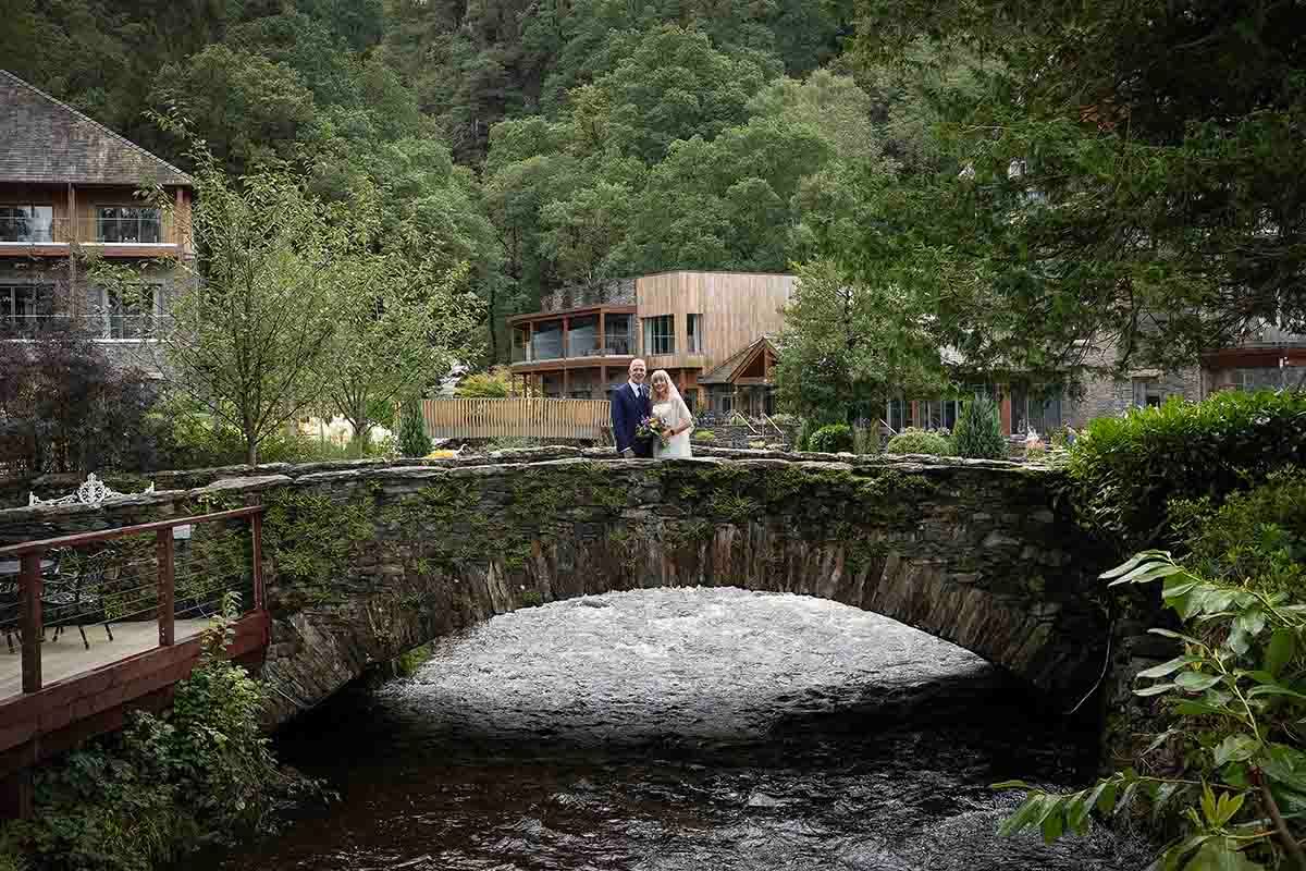 Couple embracing on a rustic stone bridge surrounded by lush greenery and a woodland venue in the background.