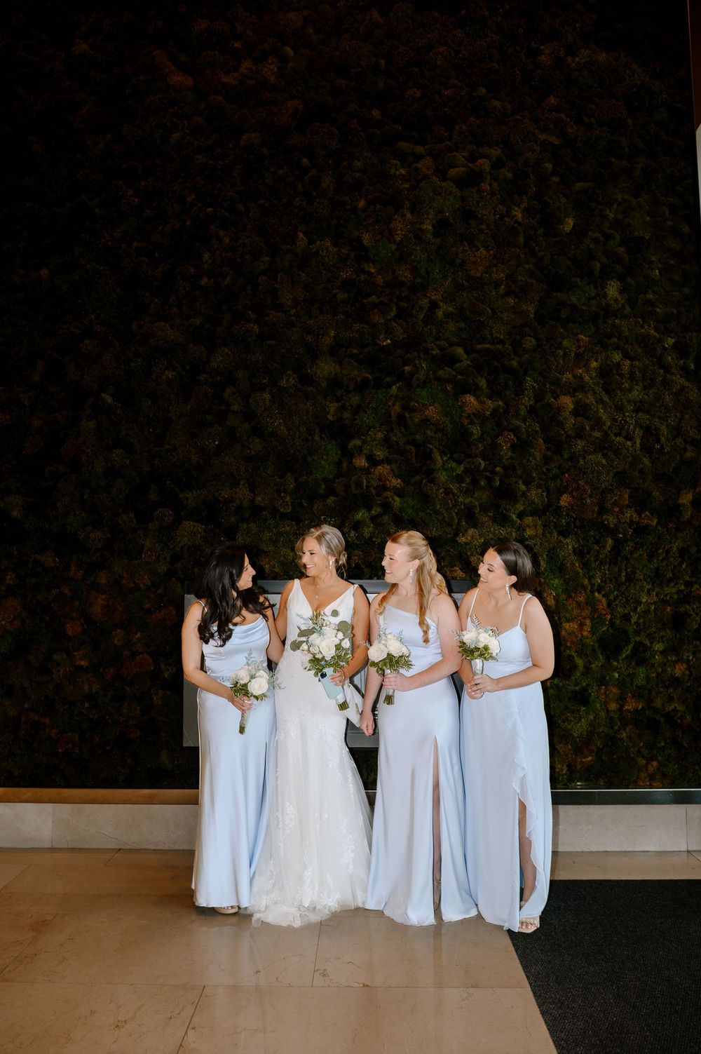 Bride in white lace gown with three bridesmaids in light blue satin dresses holding white bouquets before green wall.