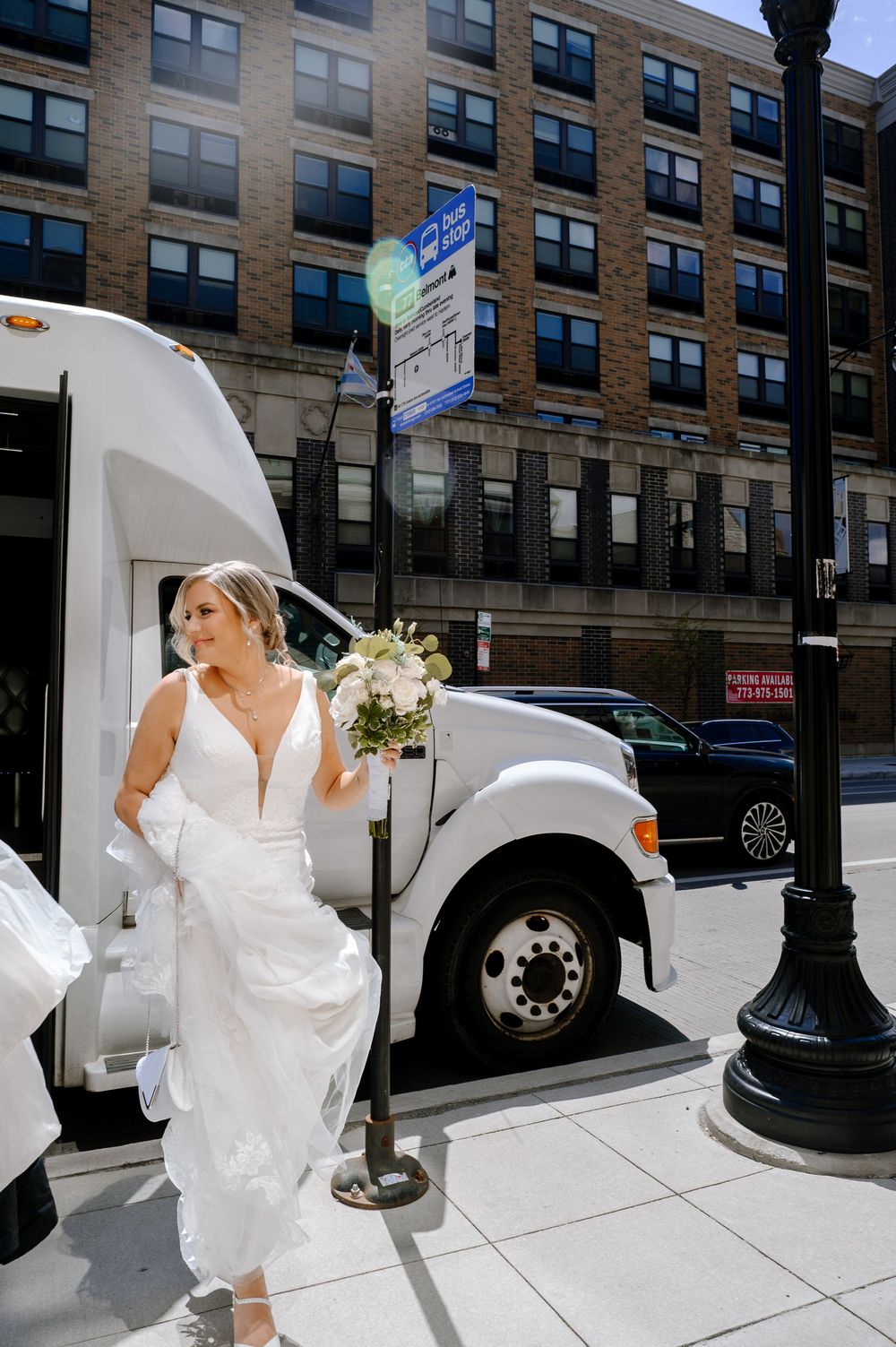 Bride in white gown holding bouquet stepping off shuttle bus on urban street on sunny wedding day.