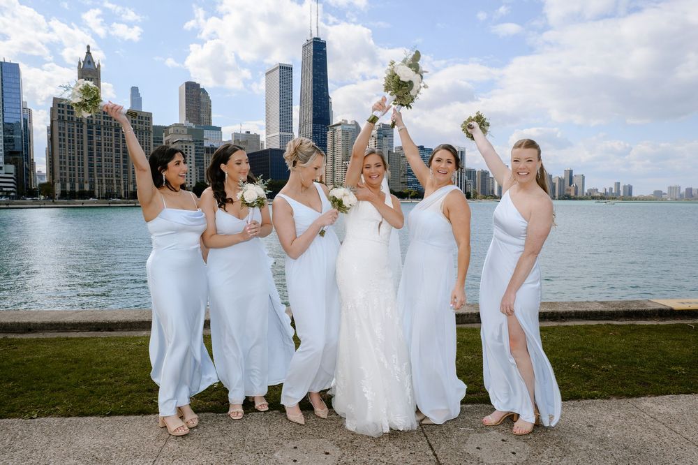 Bride and bridesmaids in white dresses celebrate with bouquets by Chicago skyline and Lake Michigan.