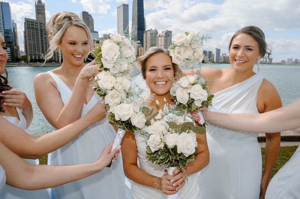 Bride and bridesmaids in white dresses holding floral bouquets with Chicago skyline in background.