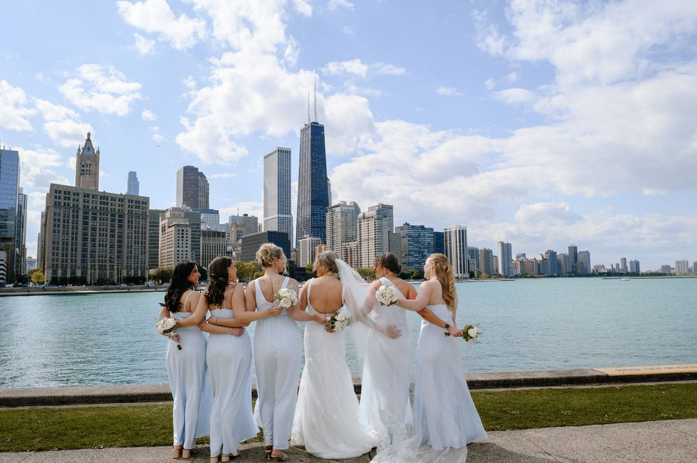 Bride and bridesmaids in white gowns pose with Chicago skyline and Lake Michigan backdrop on a sunny day.