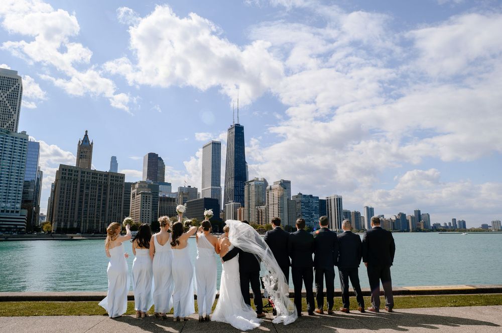 Wedding party kissing and posing by Chicago skyline waterfront with John Hancock Tower on a partly cloudy day.