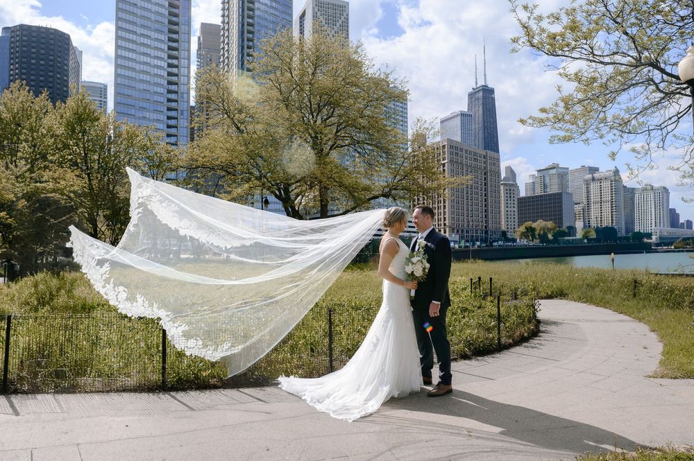 Bride and groom pose on Chicago lakefront path with veil flowing in wind, city skyline and John Hancock tower behind them.