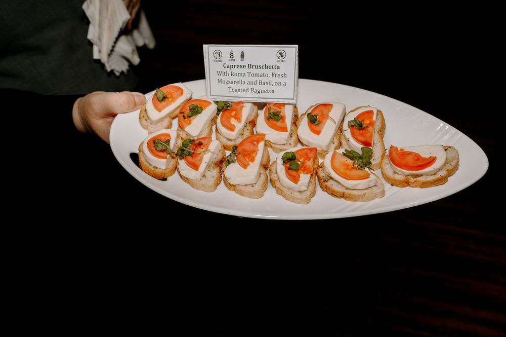 A server holds a white platter of Caprese bruschetta topped with fresh tomatoes, mozzarella, and basil at a catered event.