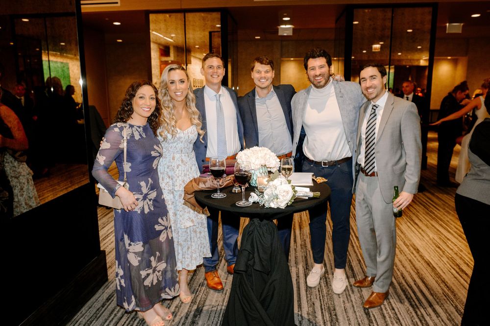 Six guests smiling and posing together at an elegant indoor event with floral centerpiece on a dark table.