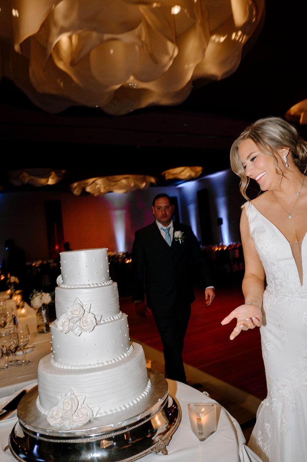 Bride in white gown smiling at elegant four-tier white wedding cake at reception venue.