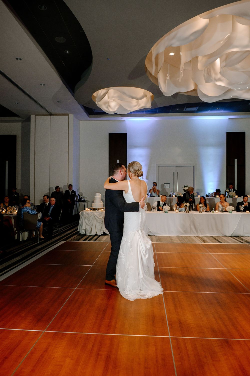 Bride and groom share their first dance on a wooden floor at an elegant wedding reception with guests seated nearby.