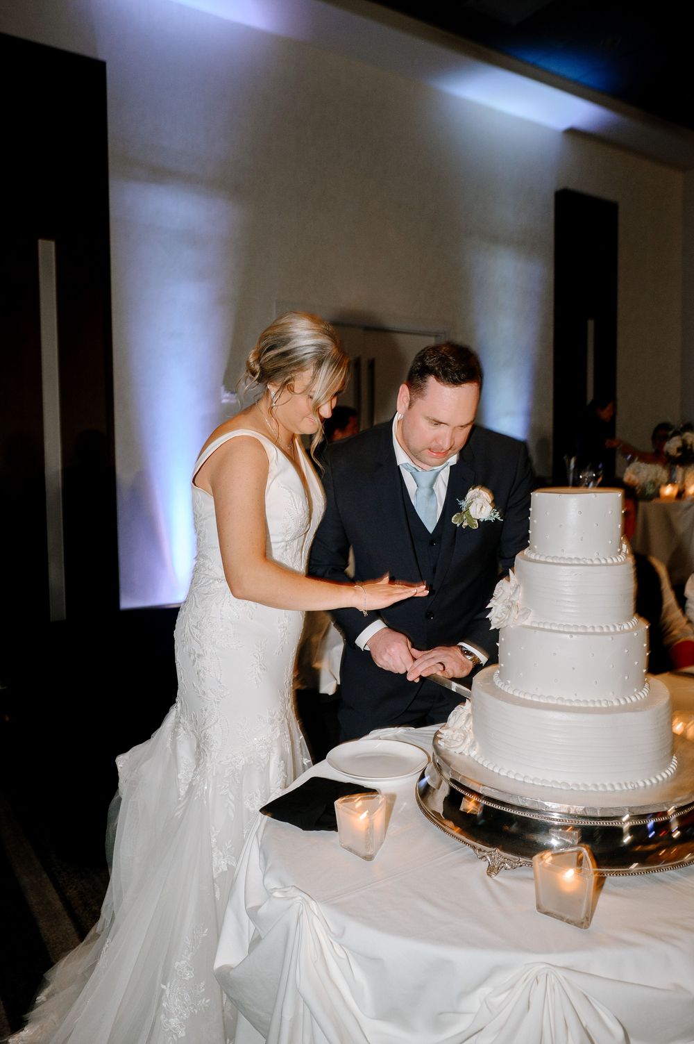 Bride and groom cutting their white tiered wedding cake at elegant reception with soft purple uplighting.
