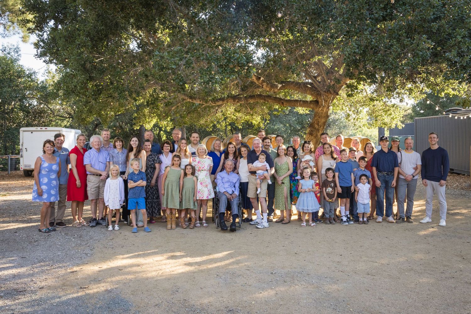 Large family group portrait outdoors under a sprawling oak tree on a sunny day.