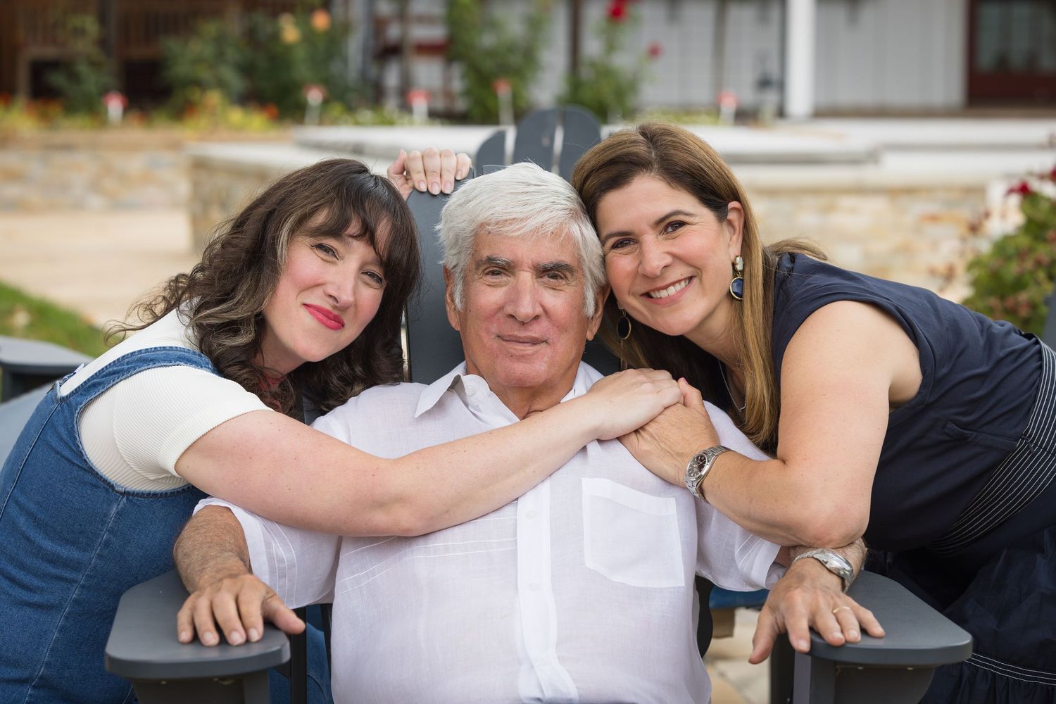 An older man smiles warmly while two women lean in close beside him outdoors.