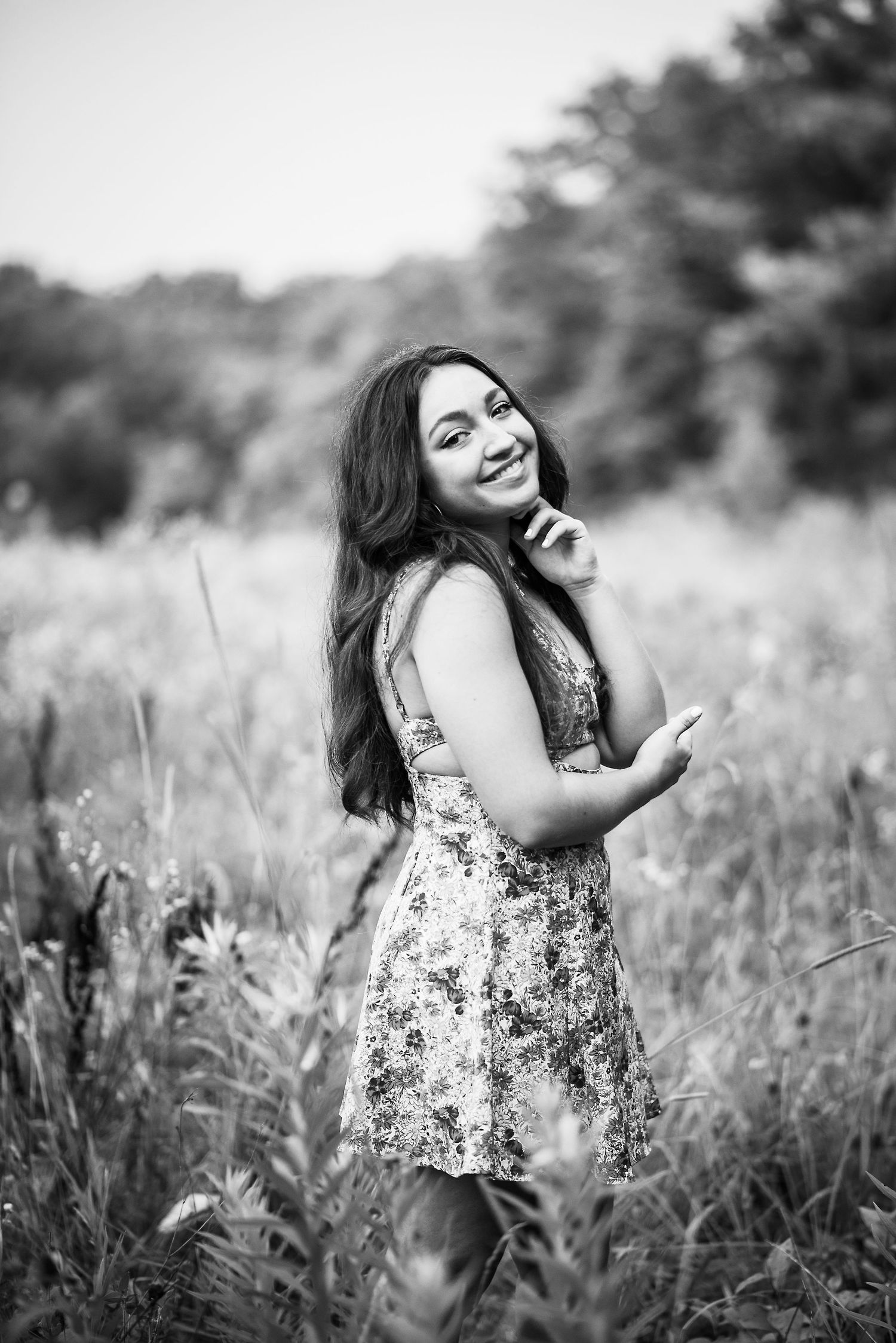 Black and white portrait of a smiling young woman in a floral dress standing in a field of tall grass.
