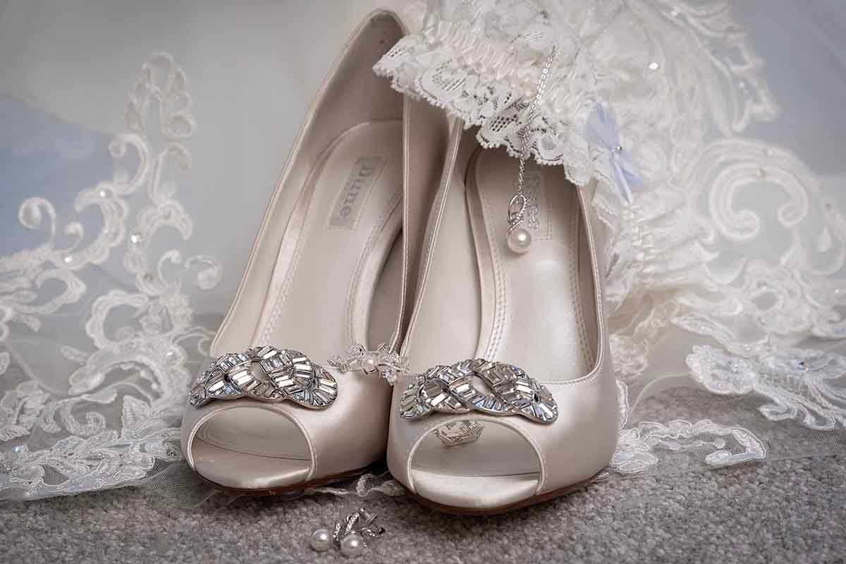 Elegant white peep-toe bridal heels with crystal embellishments displayed with lace veil and pearl earrings.