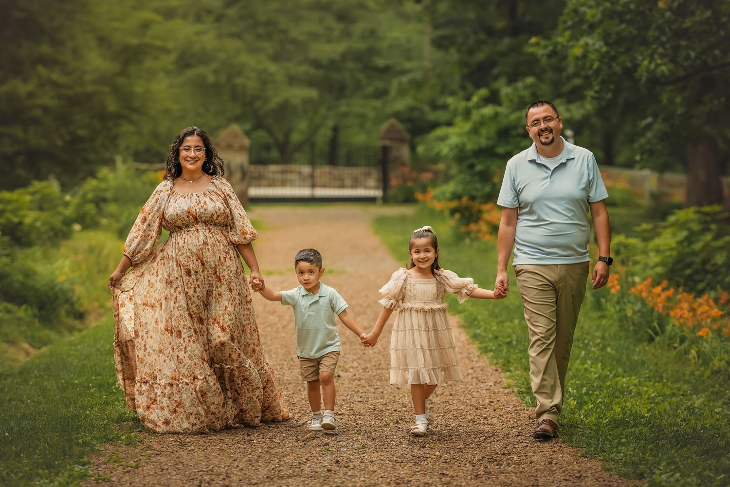 Family of four walking hand-in-hand on a garden path, parents with two young children in elegant outdoor portrait.