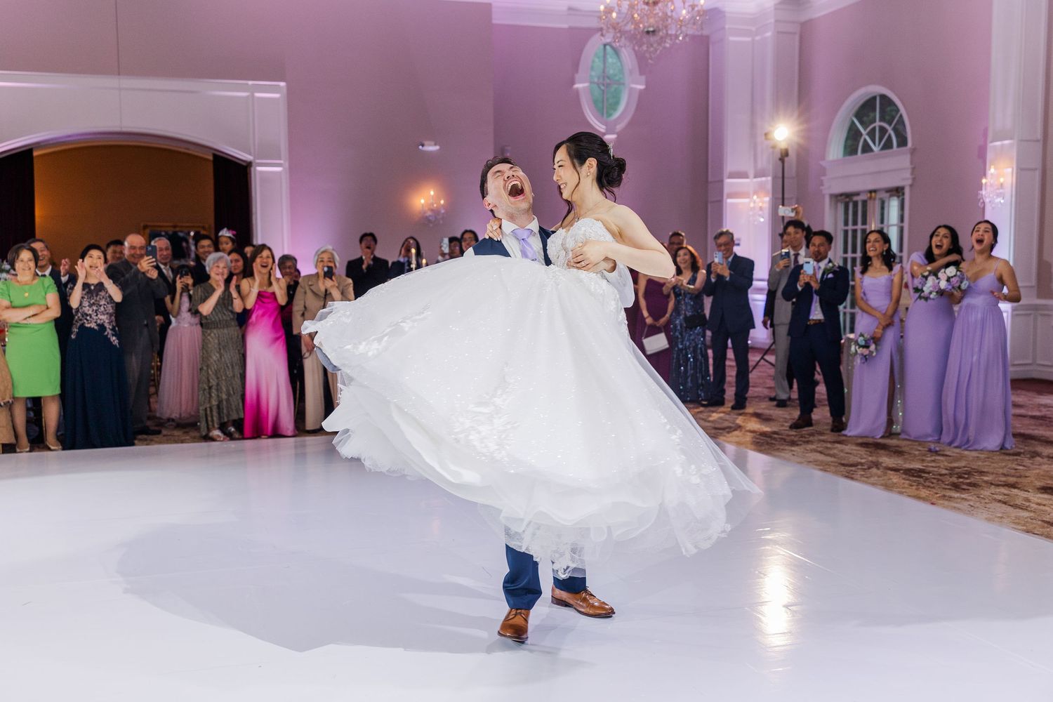 Groom lifting bride in white gown during first dance at elegant wedding reception with guests in lavender dresses.