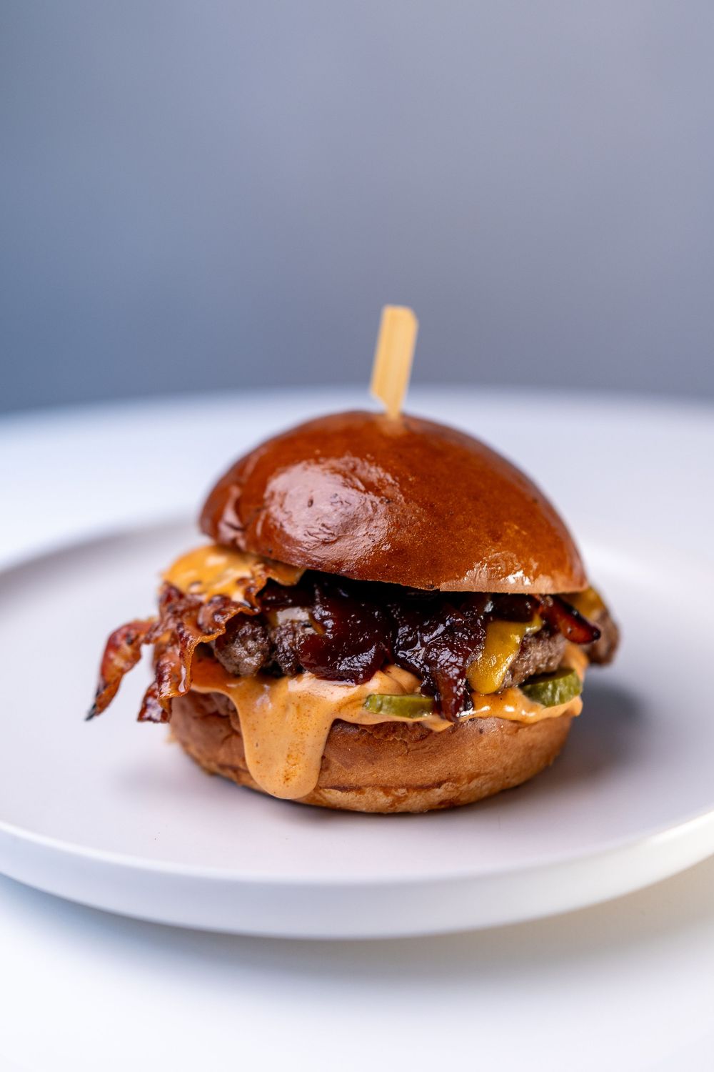 Juicy gourmet burger with melted cheese, crispy bacon, and sauce on a brioche bun, served on a white plate.