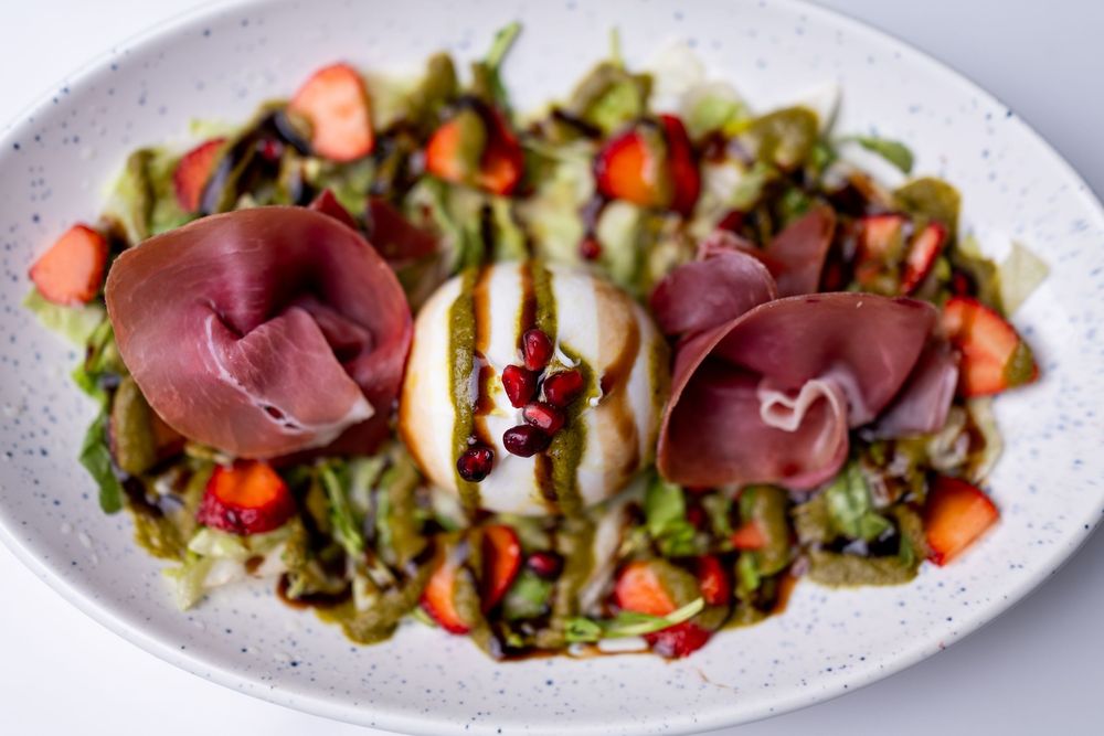 Burrata salad with prosciutto, fresh strawberries, green herbs, and balsamic glaze on a white ceramic plate.