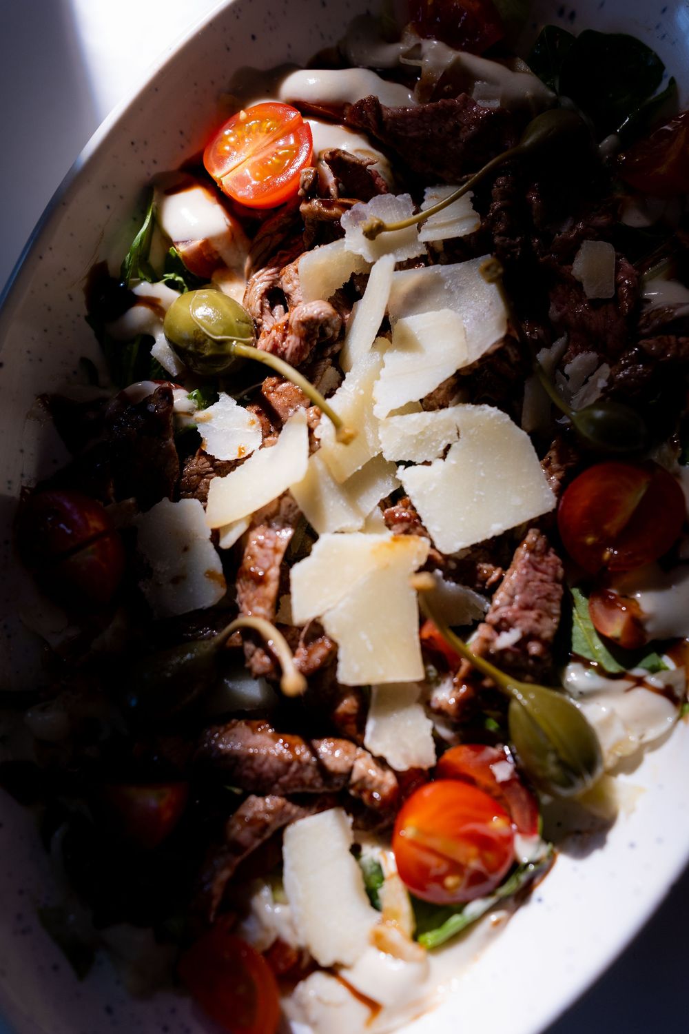 Fresh salad with grilled beef, cherry tomatoes, green olives, arugula, and shaved Parmesan cheese in a white bowl.
