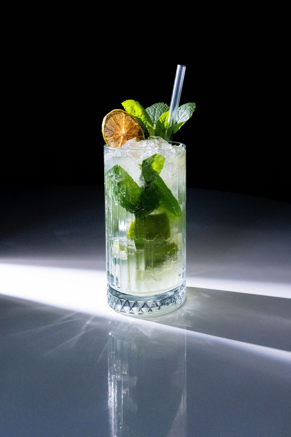 A refreshing mojito cocktail in a crystal glass with mint, lime slice, ice, and a metal straw on a reflective surface.