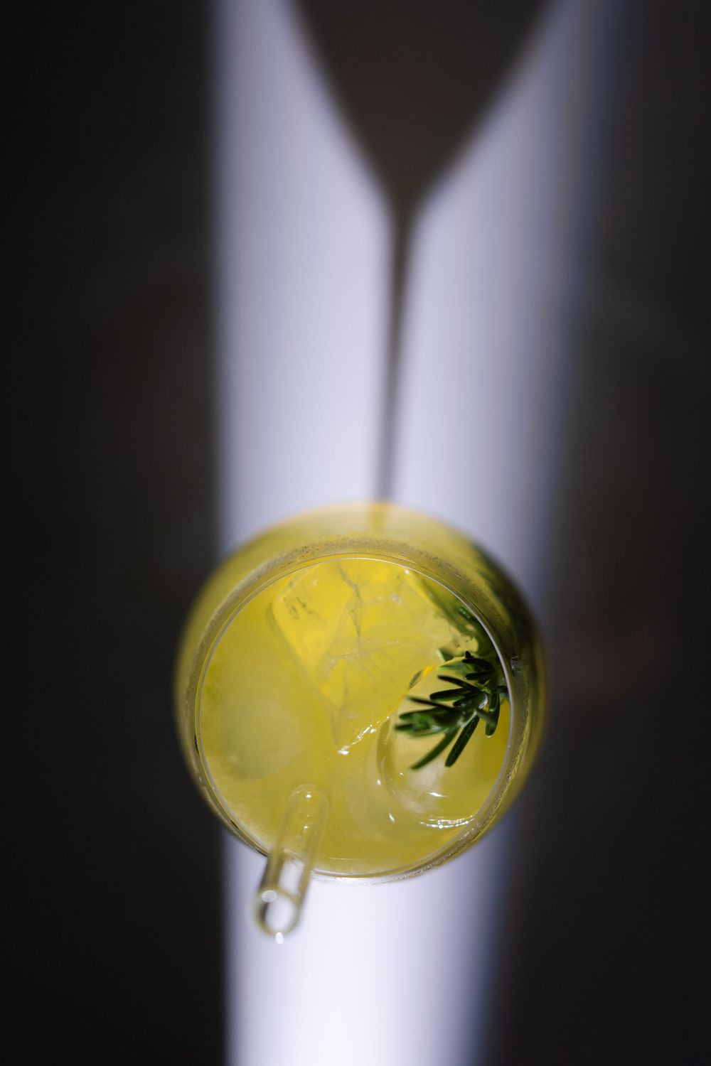 Overhead view of a yellow cocktail with ice, rosemary garnish, and dramatic light streaks on dark background.