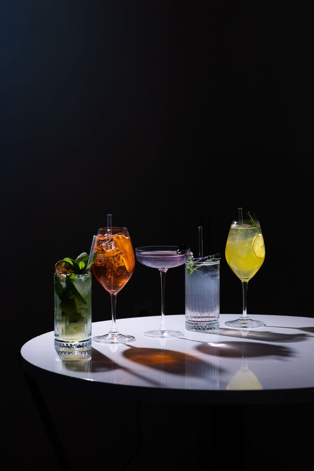 Five colorful cocktails on a white round table against a dark background, including mojito, Aperol Spritz, and citrus drinks.